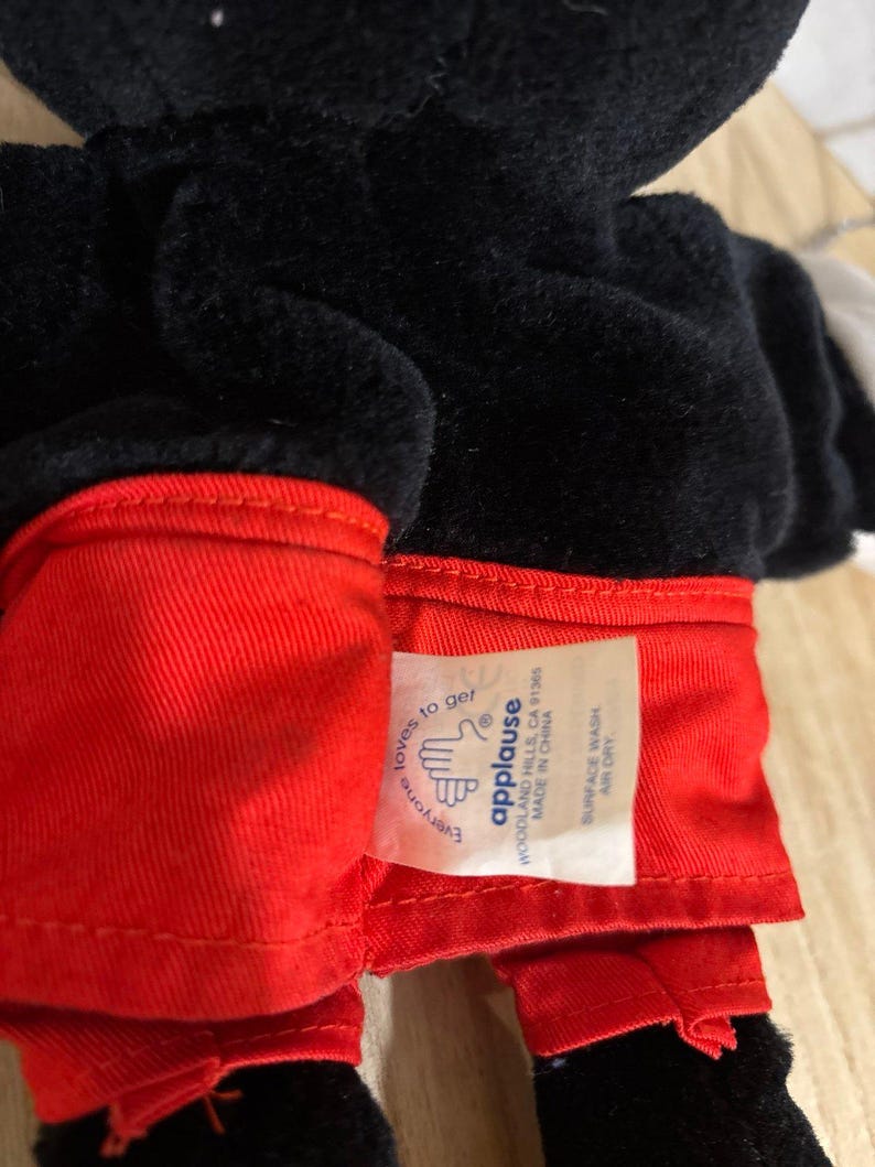 Pu&ograve; includere: Un peluche nero che indossa pantaloncini rosso brillante. Un'etichetta in tessuto bianco &egrave; attaccata alla vita, con il testo "applause", "WOODLAND HILLS, CA 91365", "MADE IN CHINA", "SURFACE WASH", "AIR DRY" e "Everyone loves to get" con un'icona a mano.