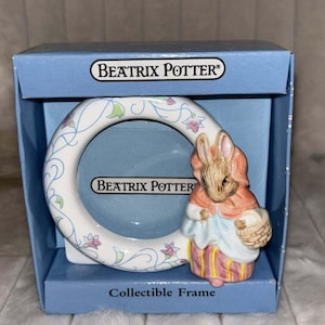May include: A round ceramic Beatrix Potter collectible frame featuring a white border with blue and pink floral vines. A three-dimensional figure of a rabbit wearing a pink shawl and striped skirt is attached to the side. The blue box displays the text Beatrix Potter.