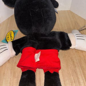 Pu&ograve; includere: Una marionetta a mano in peluche di Topolino con pelliccia nera, pantaloncini rossi e scarpe gialle. Il personaggio ha guanti bianchi con linee nere cucite. Un'etichetta laterale riporta la scritta I'M A PUPPET! in nero su sfondo giallo.