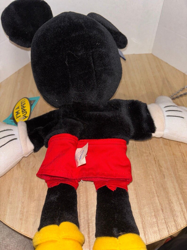 Pu&ograve; includere: Una marionetta a mano in peluche di Topolino con pelliccia nera, pantaloncini rossi e scarpe gialle. Il personaggio ha guanti bianchi con linee nere cucite. Un'etichetta laterale riporta la scritta I'M A PUPPET! in nero su sfondo giallo.