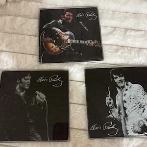 May include: Three square coasters featuring black and white photographic prints of Elvis Presley. One shows him playing a guitar, another singing into a microphone, and the third in a white jumpsuit. Each coaster includes the printed Elvis Presley signature.