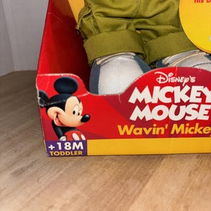 May include: A boxed Disney's Mickey Mouse toddler doll. The box is red with the words "Disney's MICKEY MOUSE Wavin' Mickey" in white and yellow. The doll is wearing green pants, a red shirt, and blue and white shoes. The box is labeled "+18M TODDLER".