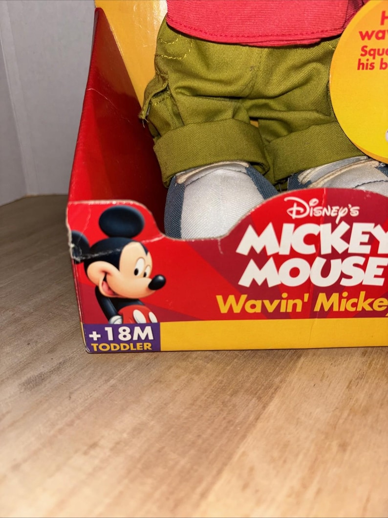 May include: A boxed Disney's Mickey Mouse toddler doll. The box is red with the words "Disney's MICKEY MOUSE Wavin' Mickey" in white and yellow. The doll is wearing green pants, a red shirt, and blue and white shoes. The box is labeled "+18M TODDLER".