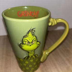 May include: A green ceramic mug featuring the Grinch. The mug has a light green exterior with a Grinch illustration and the word "GRINCH!" in red inside the rim. The base has a diamond pattern.