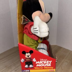 May include: A Mickey Mouse plush toy in its original packaging. The toy is wearing a red shirt, green pants, and white gloves. The packaging is red and features the words "Disney's Mickey Mouse Wavin' Mickey" and a graphic of Mickey Mouse.