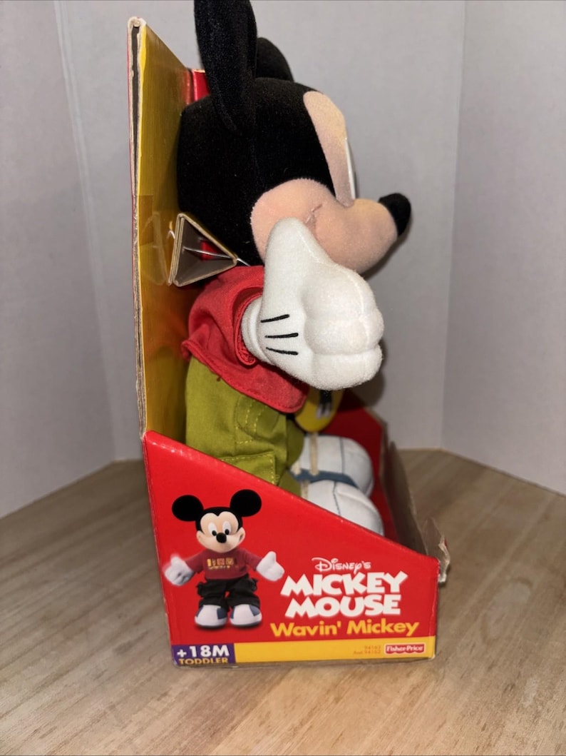May include: A Mickey Mouse plush toy in its original packaging. The toy is wearing a red shirt, green pants, and white gloves. The packaging is red and features the words "Disney's Mickey Mouse Wavin' Mickey" and a graphic of Mickey Mouse.