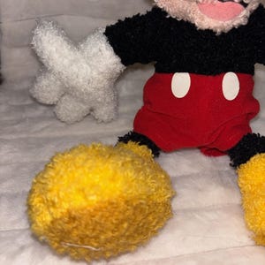 May include: A plush toy character featuring a black fuzzy torso, red shorts with two white oval buttons, white fuzzy gloves, and large yellow fuzzy shoes. The character has a light-colored fuzzy head with a visible pink mouth area.