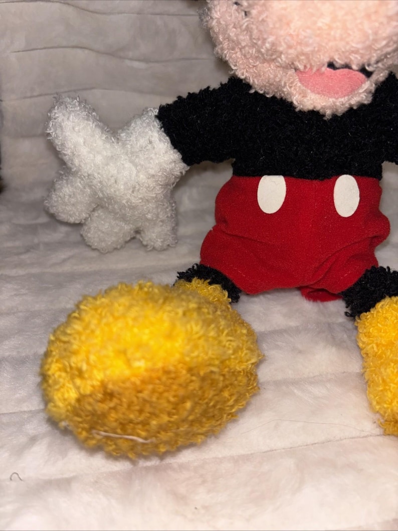 May include: A plush toy character featuring a black fuzzy torso, red shorts with two white oval buttons, white fuzzy gloves, and large yellow fuzzy shoes. The character has a light-colored fuzzy head with a visible pink mouth area.