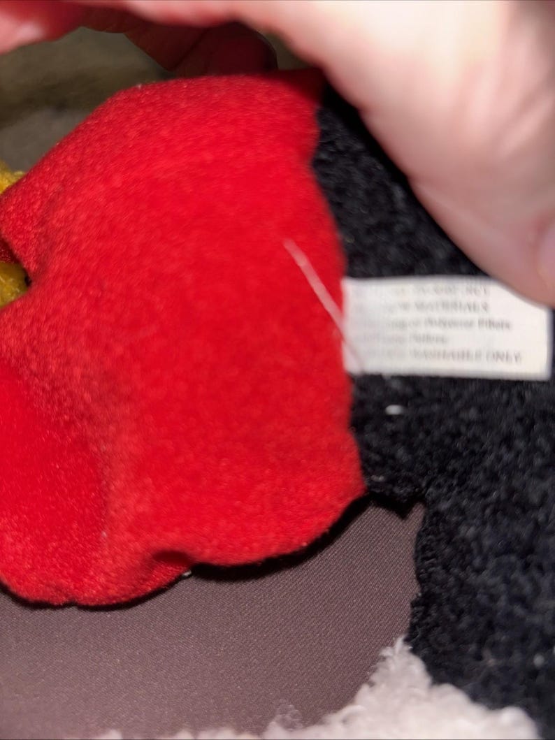 May include: A plush toy featuring a bright red fabric section adjacent to a textured black fabric area. A white rectangular fabric tag is attached to the side, displaying printed text that includes the words SURFACE WASHABLE ONLY.