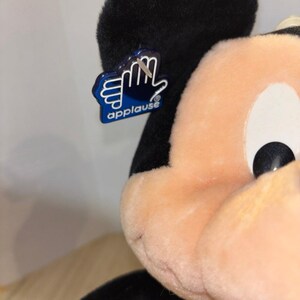 Pu&ograve; includere: Un peluche di Topolino con orecchie nere e viso color pesca. Un'etichetta in plastica blu a forma di mano &egrave; attaccata all'orecchio sinistro, con la scritta bianca "applause" e un simbolo di marchio registrato sotto le dita.