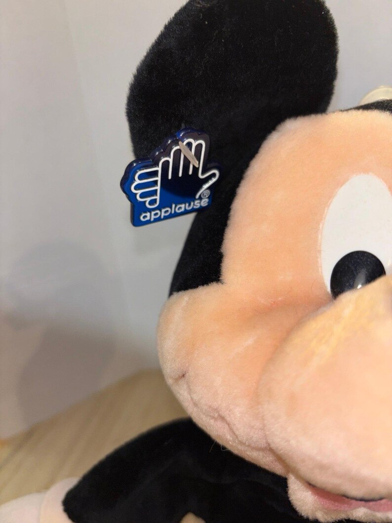 Pu&ograve; includere: Un peluche di Topolino con orecchie nere e viso color pesca. Un'etichetta in plastica blu a forma di mano &egrave; attaccata all'orecchio sinistro, con la scritta bianca "applause" e un simbolo di marchio registrato sotto le dita.