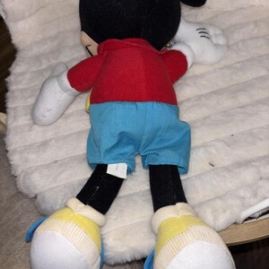 Puede incluir: Un peluche de Mickey Mouse con cabeza negra y grandes orejas redondas, camisa roja, pantalones cortos azules, guantes blancos y zapatos amarillos con detalles blancos. El personaje se muestra de espaldas, con una peque&ntilde;a etiqueta blanca en el pantal&oacute;n.