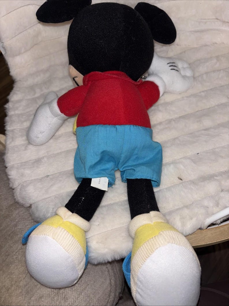 Puede incluir: Un peluche de Mickey Mouse con cabeza negra y grandes orejas redondas, camisa roja, pantalones cortos azules, guantes blancos y zapatos amarillos con detalles blancos. El personaje se muestra de espaldas, con una peque&ntilde;a etiqueta blanca en el pantal&oacute;n.