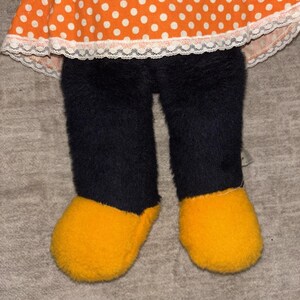 May include: A plush doll featuring an orange polka-dot dress with white lace trim, black fuzzy pants, and yellow fuzzy shoes. The doll's design is reminiscent of a cartoon character.