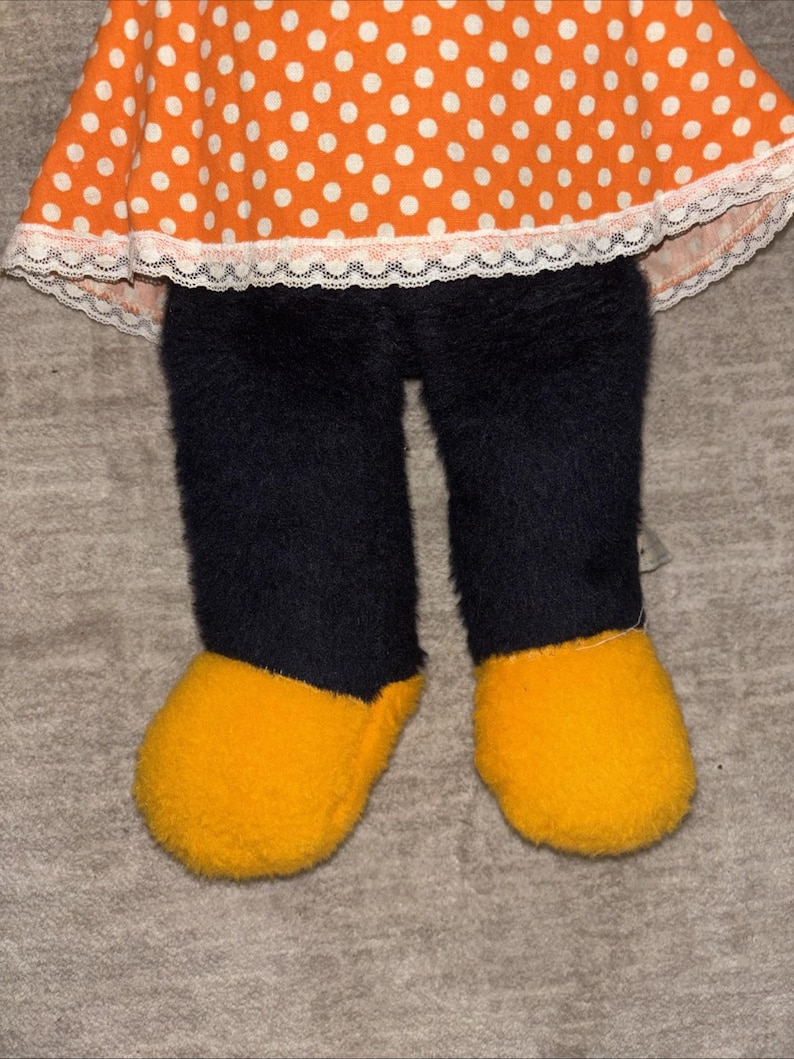 May include: A plush doll featuring an orange polka-dot dress with white lace trim, black fuzzy pants, and yellow fuzzy shoes. The doll's design is reminiscent of a cartoon character.