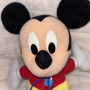 Puede incluir: Un peluche de Mickey Mouse con grandes orejas negras, cara color melocot&oacute;n y ojos ovalados blancos con pupilas negras. El personaje viste una camiseta roja de manga larga, un cintur&oacute;n amarillo con hebilla verde, pantalones azules y guantes blancos.