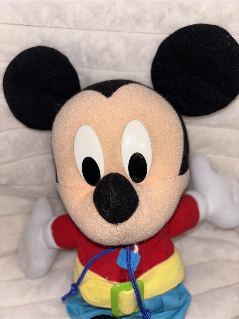 Puede incluir: Un peluche de Mickey Mouse con grandes orejas negras, cara color melocot&oacute;n y ojos ovalados blancos con pupilas negras. El personaje viste una camiseta roja de manga larga, un cintur&oacute;n amarillo con hebilla verde, pantalones azules y guantes blancos.