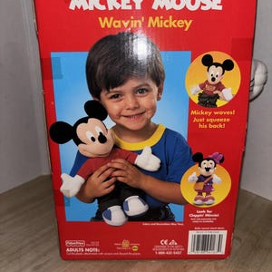 May include: A red box featuring Disney's Mickey Mouse Wavin' Mickey toy. The box has images of Mickey Mouse and a child holding the toy. Text on the box includes "Mickey waves! Just squeeze his back!" and "Look for Cloppin' Minnie!"