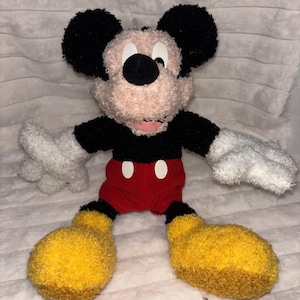 May include: A plush Mickey Mouse toy featuring a textured, fuzzy beige face, large black ears, a black shirt, red shorts with two white oval buttons, white gloves, and oversized yellow shoes. The character has a soft, fabric-like appearance.