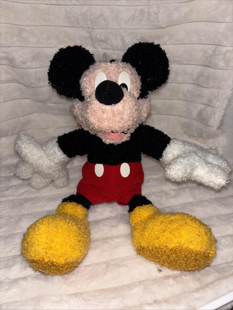May include: A plush Mickey Mouse toy featuring a textured, fuzzy beige face, large black ears, a black shirt, red shorts with two white oval buttons, white gloves, and oversized yellow shoes. The character has a soft, fabric-like appearance.