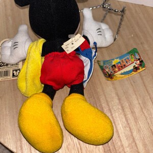 May include: A plush Mickey Mouse toy featuring a black head and body, red shorts, and large yellow shoes. The character wears white gloves and carries a yellow towel and a blue and white bag. A Florida tag and a price tag are attached to the toy.