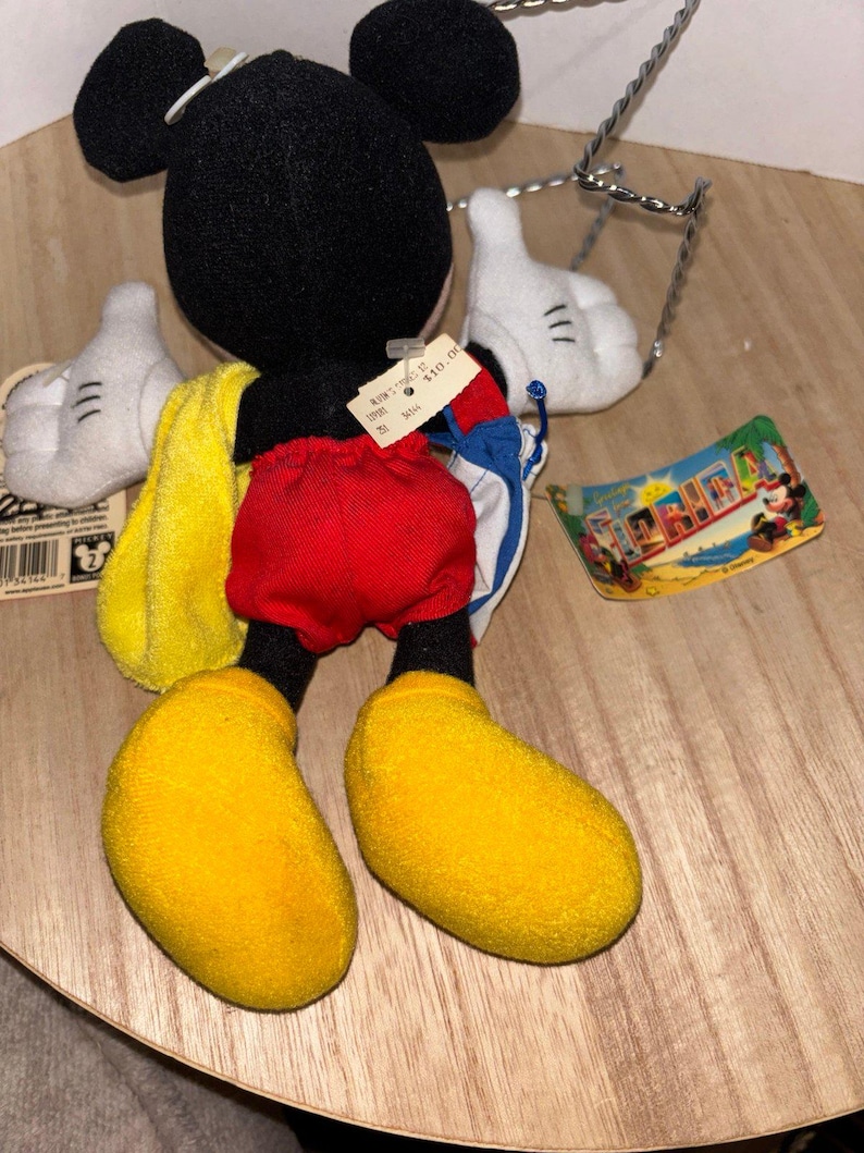 May include: A plush Mickey Mouse toy featuring a black head and body, red shorts, and large yellow shoes. The character wears white gloves and carries a yellow towel and a blue and white bag. A Florida tag and a price tag are attached to the toy.