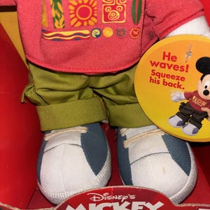May include: A Mickey Mouse plush doll wearing a red shirt with colorful graphics, green pants, and blue and white sneakers. The doll is in a red box with the text "Disney's Mickey Mouse Wavin' Mickey". A tag says "He waves! Squeeze his back."