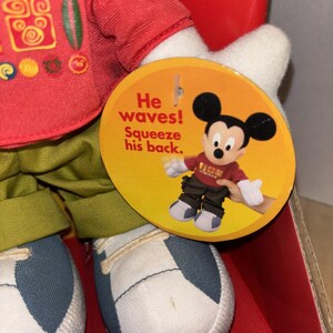 May include: A Mickey Mouse plush toy wearing a red shirt with decorative symbols, green pants, and white and blue sneakers. A round tag says "He waves! Squeeze his back." The toy is in a red box.