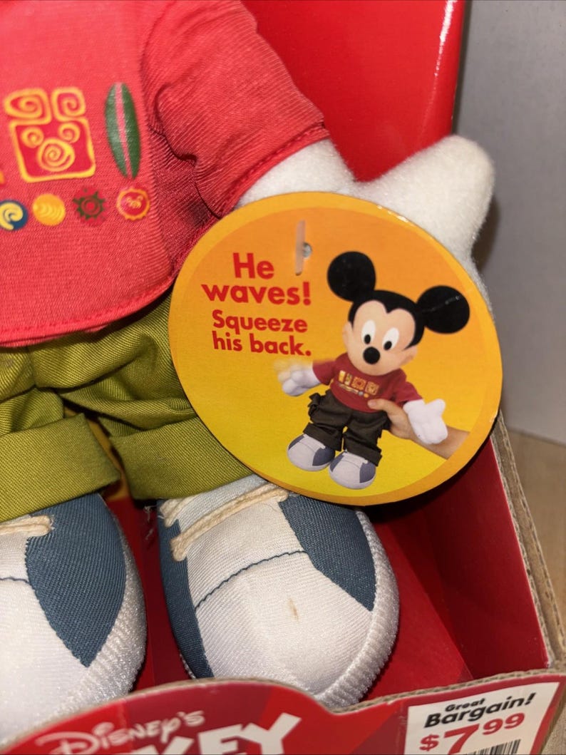 May include: A Mickey Mouse plush toy wearing a red shirt with decorative symbols, green pants, and white and blue sneakers. A round tag says "He waves! Squeeze his back." The toy is in a red box.