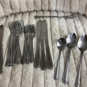 May include: A collection of stainless steel flatware including three dinner knives, eight forks, three table knives, and four spoons. Each piece features a simple, smooth handle design with a subtle beaded border detail along the edges of the stems.