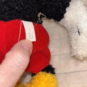 May include: A plush toy featuring a black textured torso, white textured hands, red shorts, and yellow textured feet. A white fabric tag is attached to the red shorts, displaying the text Mattel, Inc., El Segundo, CA 90245, MADE IN CHINA, and Disney.