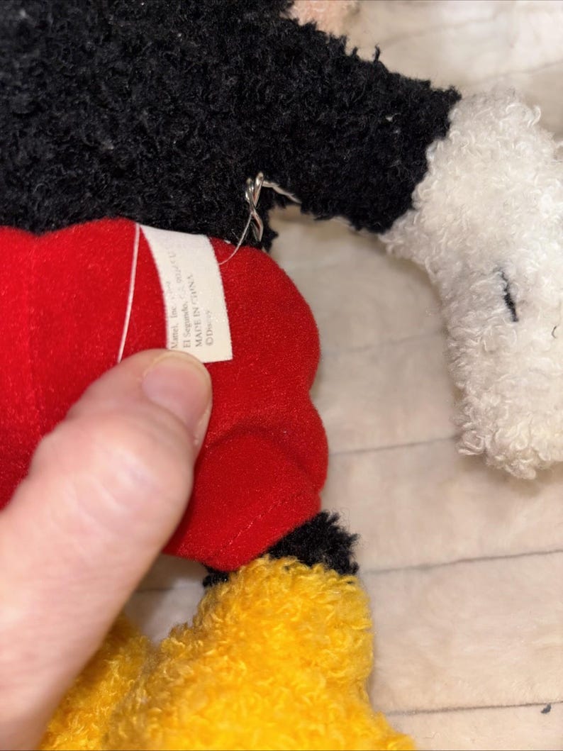 May include: A plush toy featuring a black textured torso, white textured hands, red shorts, and yellow textured feet. A white fabric tag is attached to the red shorts, displaying the text Mattel, Inc., El Segundo, CA 90245, MADE IN CHINA, and Disney.