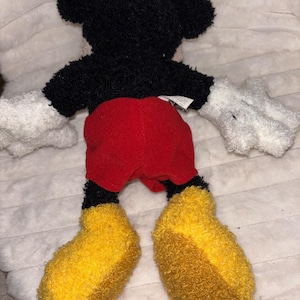 May include: A plush toy of a cartoon mouse with a black fuzzy head and torso, white fuzzy hands, bright red shorts, and large yellow fuzzy shoes. The character is shown from the back, featuring a small white fabric tag attached to the waist area.