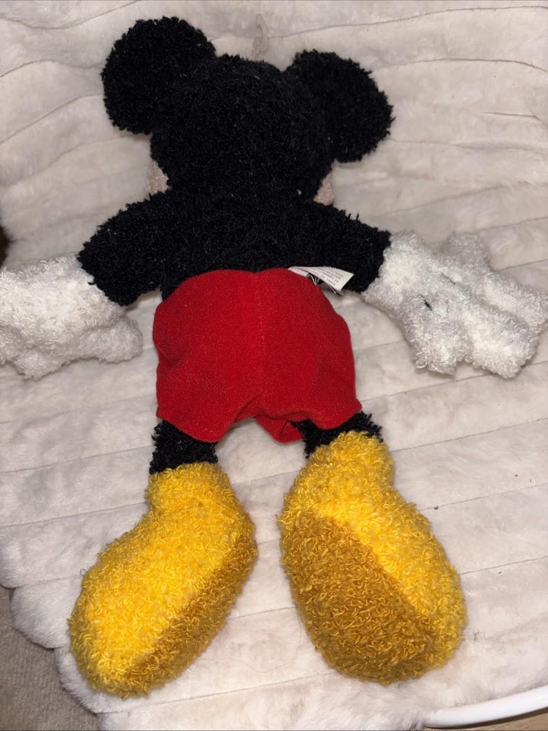 May include: A plush toy of a cartoon mouse with a black fuzzy head and torso, white fuzzy hands, bright red shorts, and large yellow fuzzy shoes. The character is shown from the back, featuring a small white fabric tag attached to the waist area.
