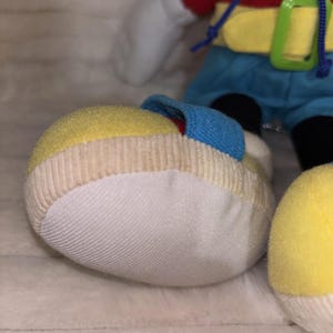Puede incluir: Un peluche con un zapato grande y redondeado de color amarillo y blanco con una correa azul. El juguete lleva una camisa roja, un cintur&oacute;n amarillo brillante con una hebilla verde y pantalones cortos azules. Un cord&oacute;n azul cuelga de la camisa y se ven manos blancas.