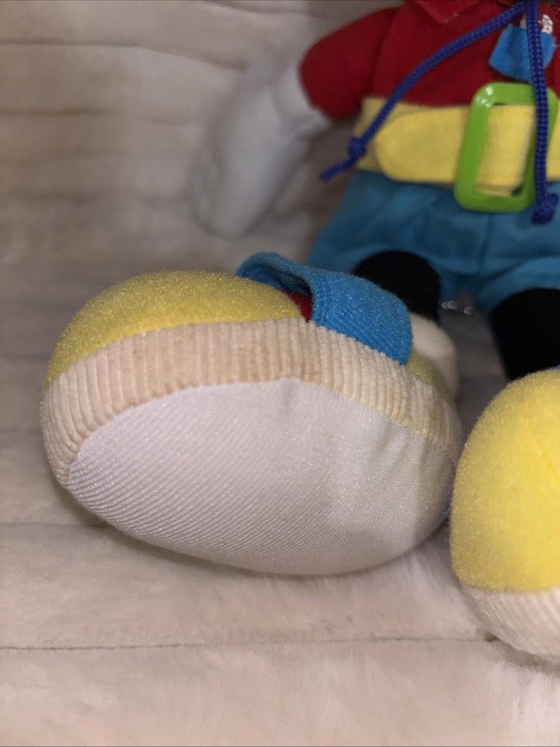 Puede incluir: Un peluche con un zapato grande y redondeado de color amarillo y blanco con una correa azul. El juguete lleva una camisa roja, un cintur&oacute;n amarillo brillante con una hebilla verde y pantalones cortos azules. Un cord&oacute;n azul cuelga de la camisa y se ven manos blancas.