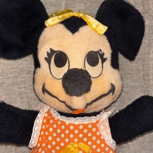May include: A vintage Minnie Mouse plush toy. The plush features black ears, a yellow bow, and a smiling face. She wears an orange dress with white polka dots and a yellow bow at the waist. The toy has black arms and is set against a neutral background.