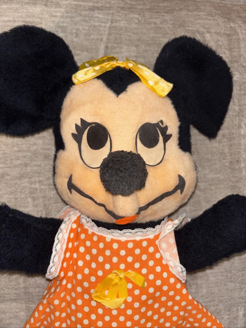 May include: A vintage Minnie Mouse plush toy. The plush features black ears, a yellow bow, and a smiling face. She wears an orange dress with white polka dots and a yellow bow at the waist. The toy has black arms and is set against a neutral background.