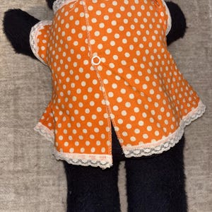 May include: A black plush toy wearing an orange dress with white polka dots. The dress has white lace trim and a button closure down the back. The toy's feet are yellow.