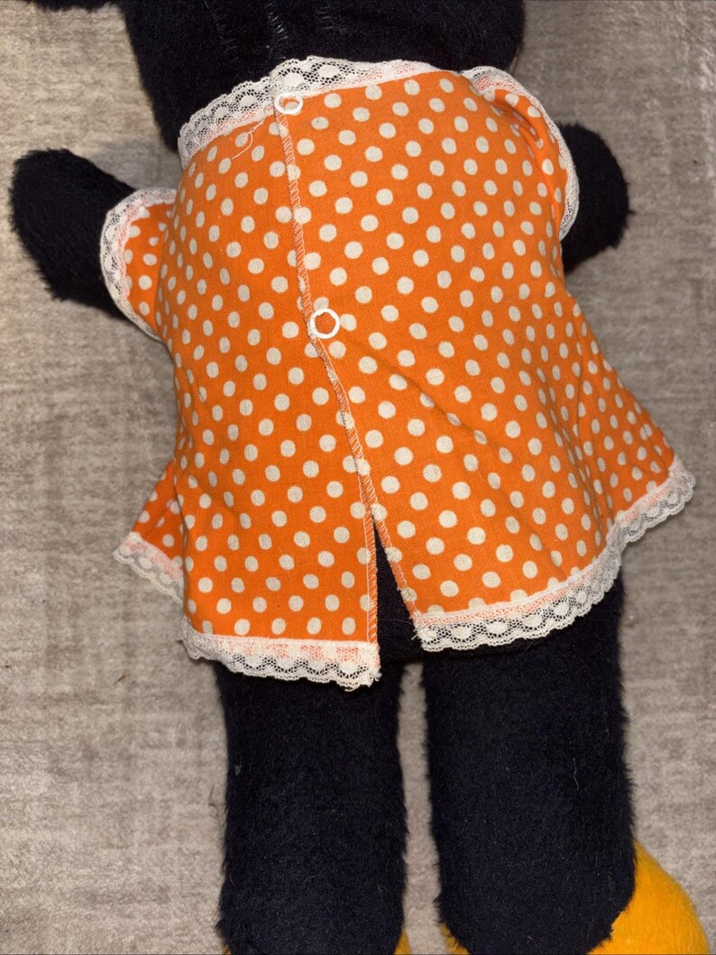 May include: A black plush toy wearing an orange dress with white polka dots. The dress has white lace trim and a button closure down the back. The toy's feet are yellow.