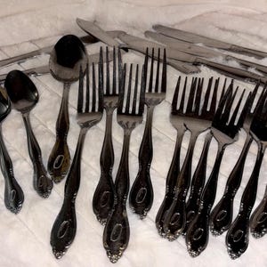 May include: A collection of dark metallic flatware including four spoons, four forks, and three knives. Each piece features a polished finish and a decorative oval detail at the base of the handle. The set is arranged in a group on a white surface.