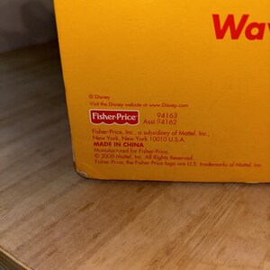 May include: Close-up of a yellow box with the Fisher-Price logo and Disney branding. Text includes "MADE IN CHINA" and a website address. The box is on a wooden surface.