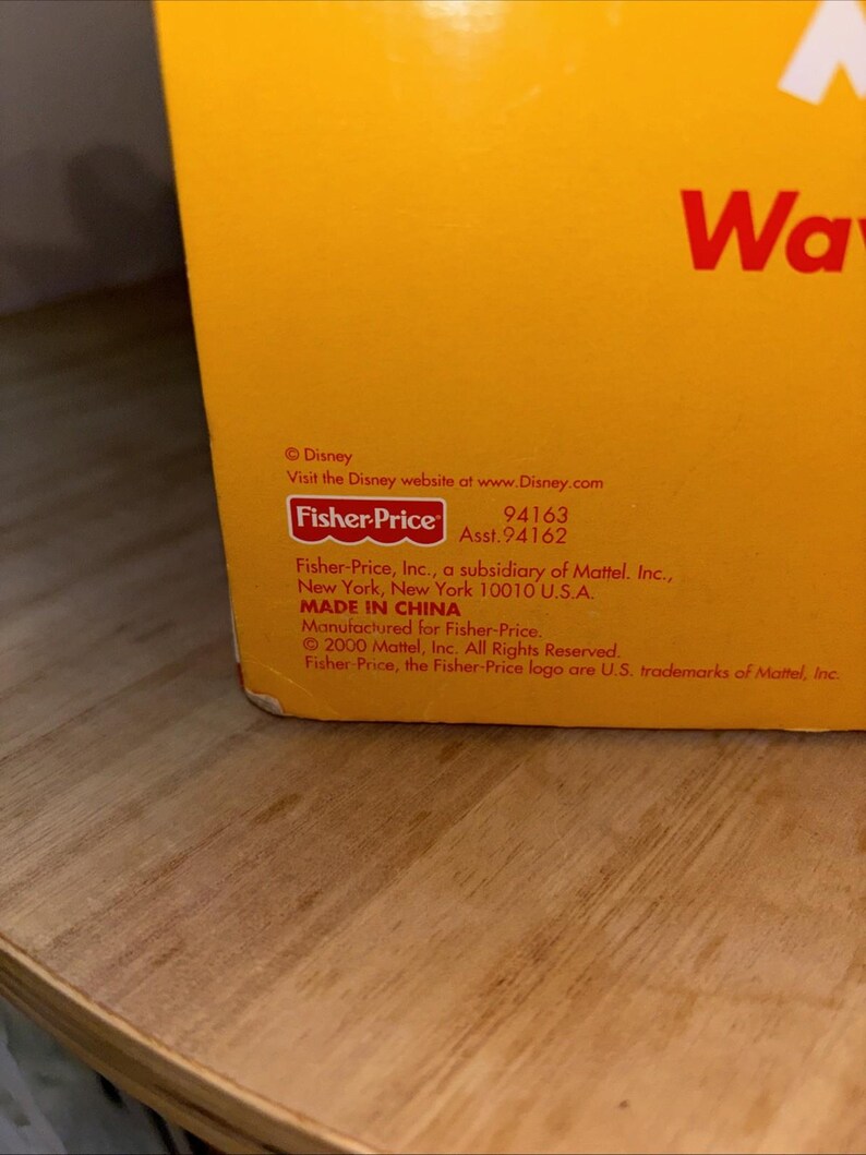 May include: Close-up of a yellow box with the Fisher-Price logo and Disney branding. Text includes "MADE IN CHINA" and a website address. The box is on a wooden surface.
