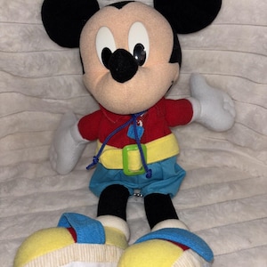 Puede incluir: Un peluche de Mickey Mouse con grandes orejas negras, cara color canela y nariz negra. El personaje viste una camisa roja con cord&oacute;n azul, un cintur&oacute;n amarillo con hebilla verde, pantalones cortos azules y zapatos grandes amarillos y azules.