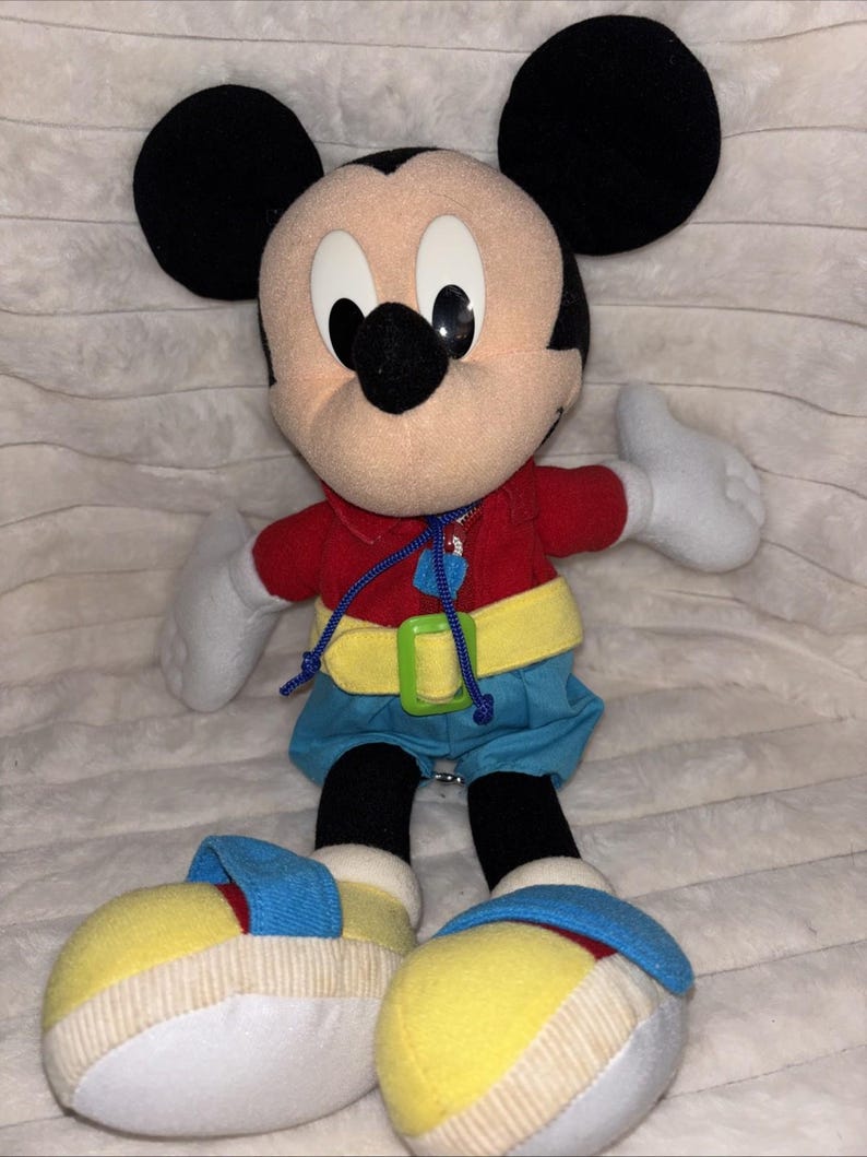 Puede incluir: Un peluche de Mickey Mouse con grandes orejas negras, cara color canela y nariz negra. El personaje viste una camisa roja con cord&oacute;n azul, un cintur&oacute;n amarillo con hebilla verde, pantalones cortos azules y zapatos grandes amarillos y azules.