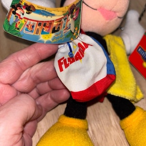 May include: A plush Mickey Mouse toy wearing a yellow cape and carrying a white, red, and blue bag with the word FLORIDA printed in red. A curved cardboard tag attached to the bag features the text Greetings from FLORIDA and a Disney logo.