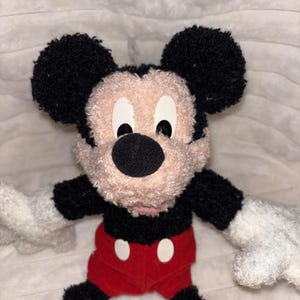 May include: A plush toy of Mickey Mouse featuring a textured, curly black and beige fabric. The character has large round black ears, a black nose, white gloves, red shorts with two white oval buttons, and bright yellow shoes on its feet.
