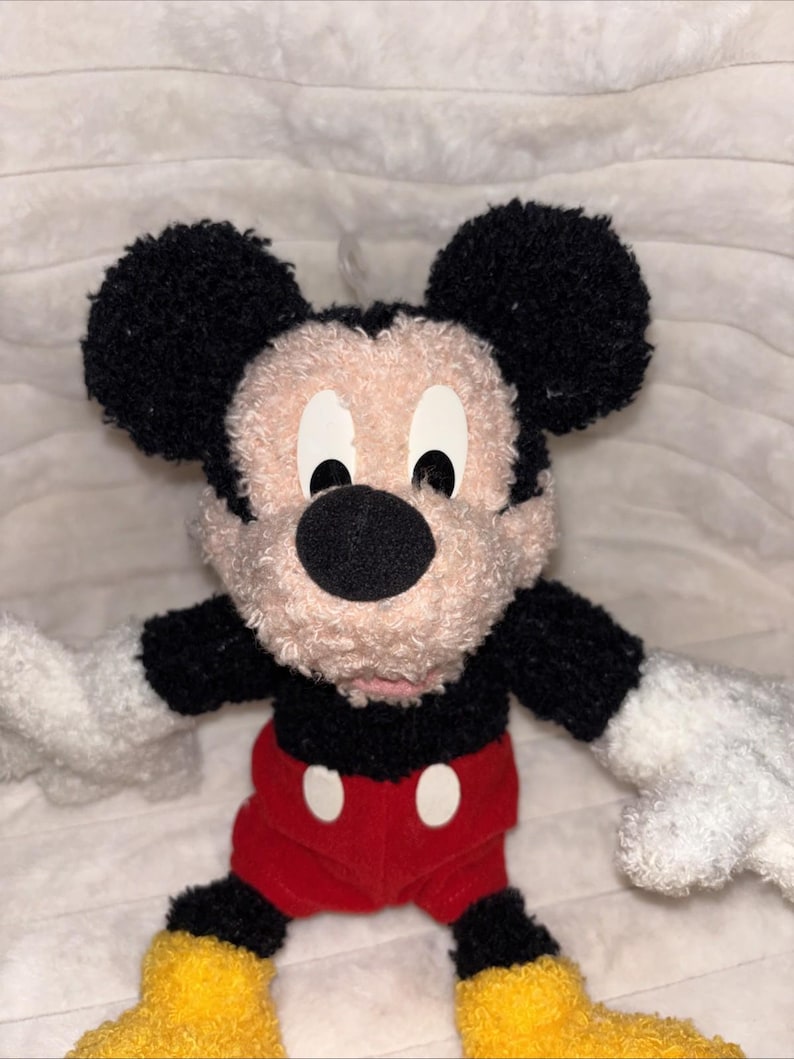 May include: A plush toy of Mickey Mouse featuring a textured, curly black and beige fabric. The character has large round black ears, a black nose, white gloves, red shorts with two white oval buttons, and bright yellow shoes on its feet.