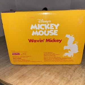 May include: A yellow box featuring the Disney's Mickey Mouse logo and the text "Wavin' Mickey" in white and red. The box includes text about the product's materials and manufacturer, Fisher-Price.