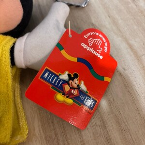 May include: A plush Mickey Mouse toy wearing a yellow garment, featuring a red cardboard tag attached by a plastic fastener. The tag displays the text "Everyone loves to get applause" and "MICKEY FOR KIDS" with a graphic of Mickey Mouse.